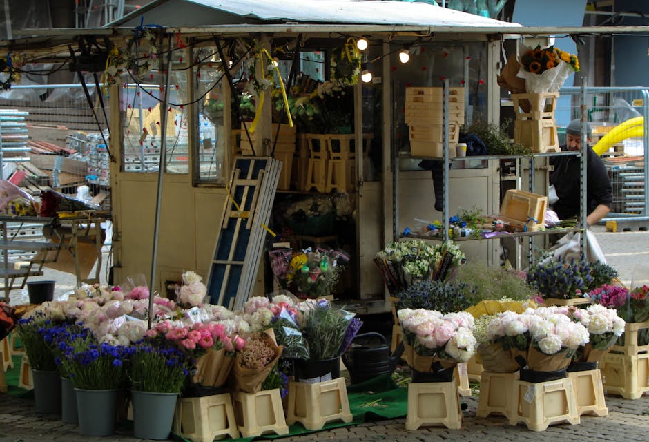 An outdoor flower stall on Golders Green Road NW11 displays a variety of fresh flowers arranged in pots and bouquets, including pink, white, and purple blooms such as roses, carnations, and chrysanthe
