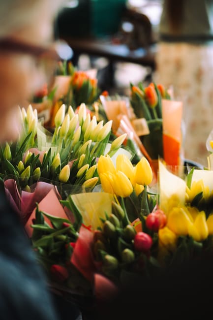 A vibrant display of freshly cut tulips in shades of yellow, white, pink, and orange, arranged in bouquets with green stems and leaves. The flowers are wrapped in pink and yellow tissue paper, with so