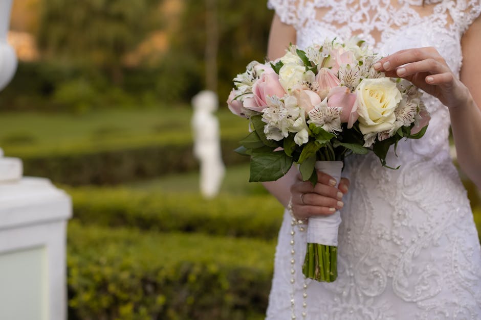 A bride wearing a white lace wedding dress holding a bouquet arrangement of fresh flowers, including pale pink roses, white roses, and white alstroemeria with pink streaks, accented by green foliage. 
