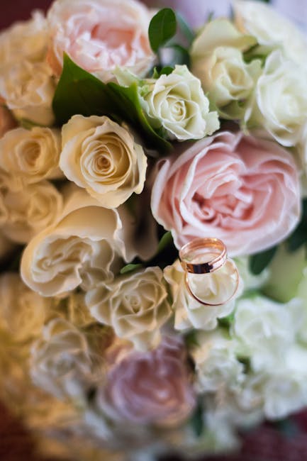 A close-up view of a floral bouquet featuring predominantly cream and soft pink roses with delicate, layered petals and lush green leaves, arranged in a dense cluster. Prominently displayed are two we
