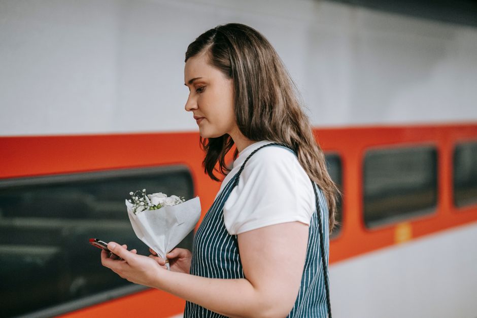 A young woman with long, wavy brown hair stands on a train platform holding a small bouquet of fresh white flowers, wrapped in white paper with green foliage peeking through. She is wearing a white sh