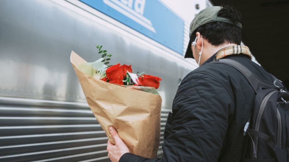 A person wearing a dark jacket, beige baseball cap, and white earphones, holding a bouquet of red roses wrapped in brown kraft paper, standing next to a silver train carriage with horizontal ridges, u