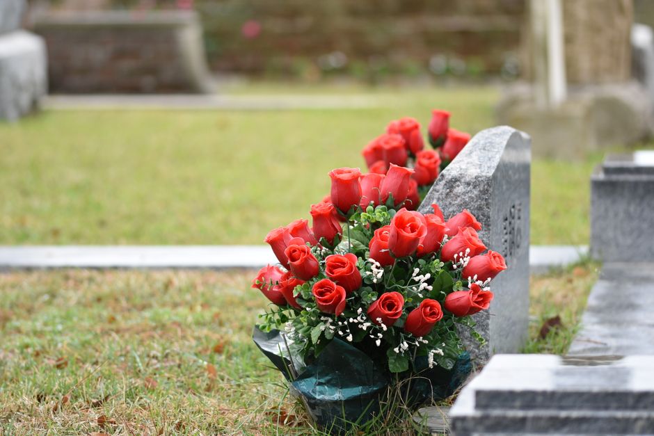 A bouquet of fresh, vibrant red roses with tightly closed petals and glossy green leaves, arranged in a compact cluster. The bouquet is wrapped in dark green floral paper and is placed at the base of 
