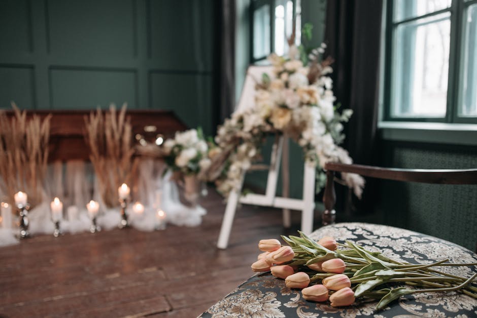 A floral arrangement scene featuring pale pink tulips with long green stems and leaves resting on a cushioned, patterned chair in the foreground. In the background, a large, elegant floral tribute com